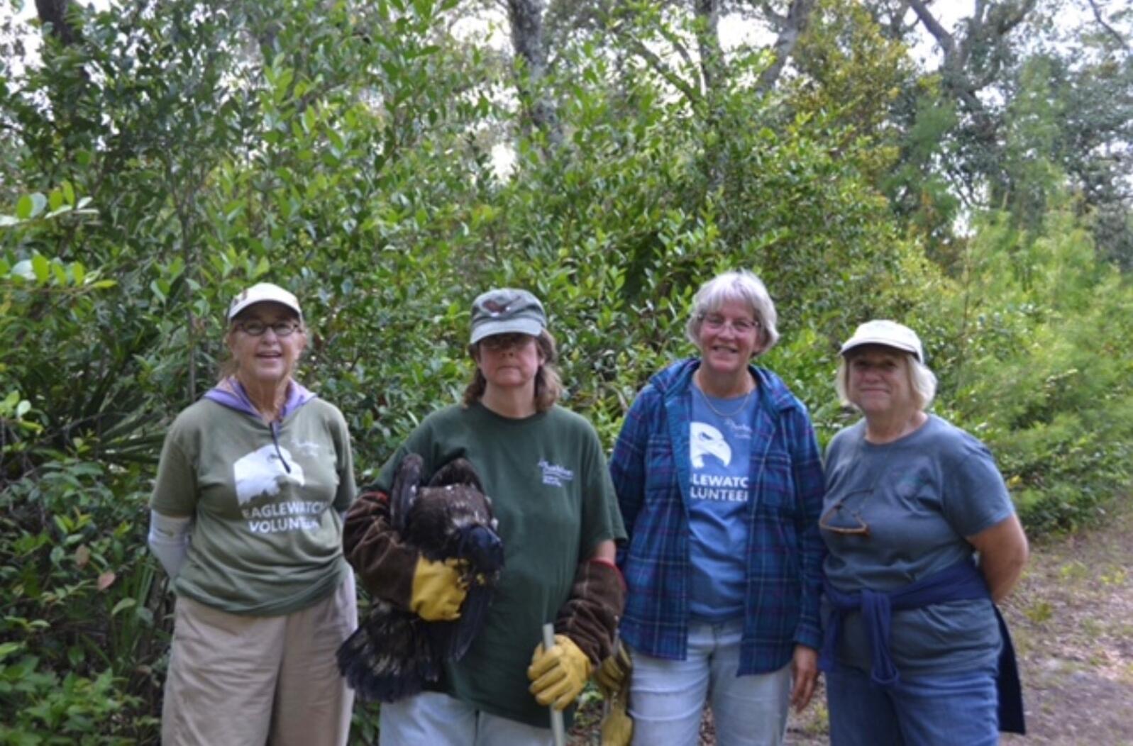 Four women stand in a line and pose for a photo in the woods. The second woman from the left is holding a hooded Bald Eagle under one arm.