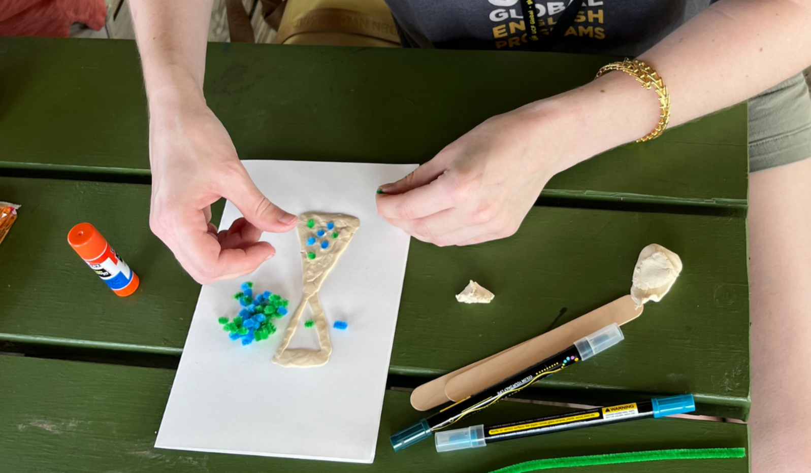 A close-up view of a student's hands placing tiny pieces of blue and green pipe cleaners into taped triangles on a piece of paper.