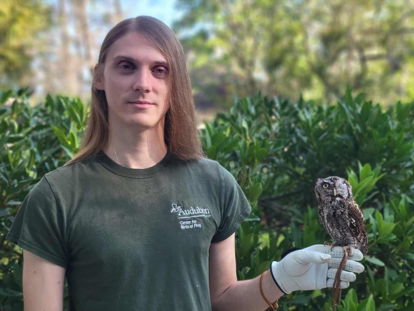 A man stands outdoors holding a small owl on his gloved hand.