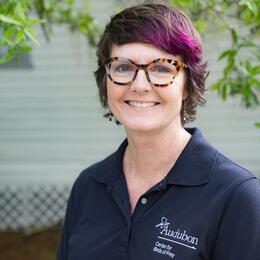 A portrait of a woman with short brown and pink hair, wearing glasses and a black polo with the Audubon Center for Birds of Prey logo.