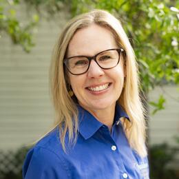 A portrait of a woman with blonde hair wearing glasses and a royal blue oxford shirt.