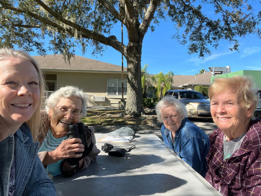 Four women sit at a picnic table, smiling at the camera. One of the women holds a pair of binoculars.